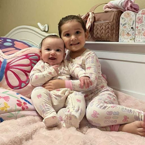 Two children in matching pajamas sitting on a bed with a decorative pillow.